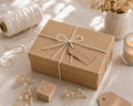 Une boîte-cadeau « Pure M » rectangulaire de couleur marron, fabriquée par Artoz Papier AG, fermée par une ficelle blanche et portant l'inscription « For You », repose sur une surface beige, entourée de fleurs séchées, d'une bougie, de ficelle et d'un tampon en bois en forme de cœur. La lumière chaude du soleil inonde la pièce.