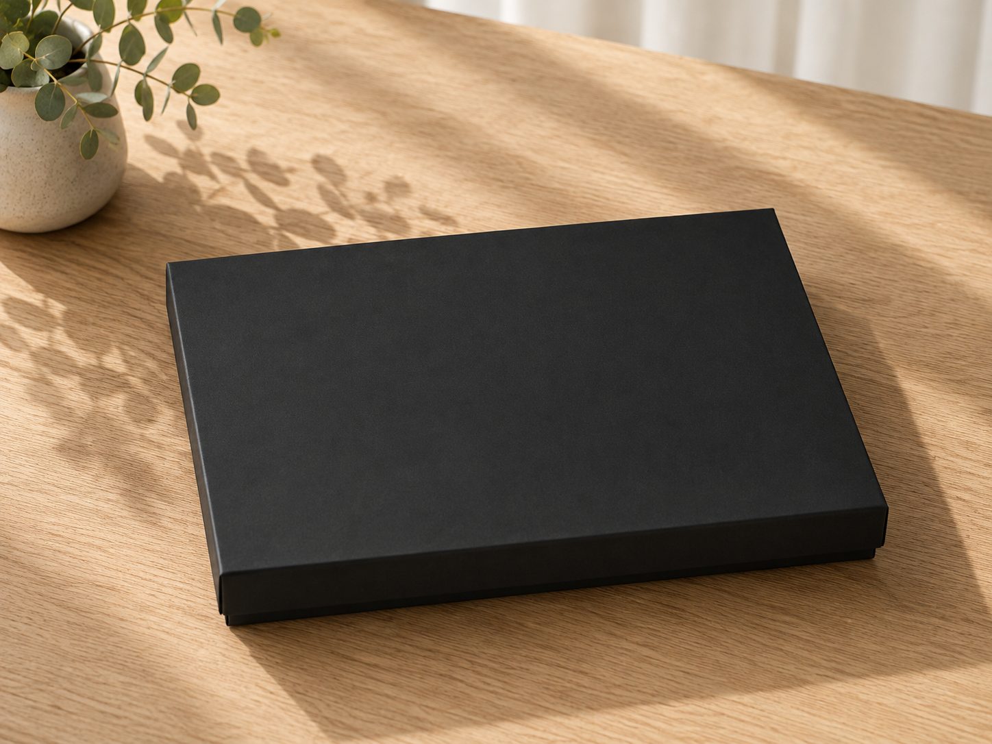 Eine geschlossene, mattschwarze Geschenkbox „Pure A4“ der Artoz Papier AG steht auf einem Holztisch neben einer kleinen Topfpflanze, wobei das Sonnenlicht sanfte Schatten auf die Oberfläche wirft.