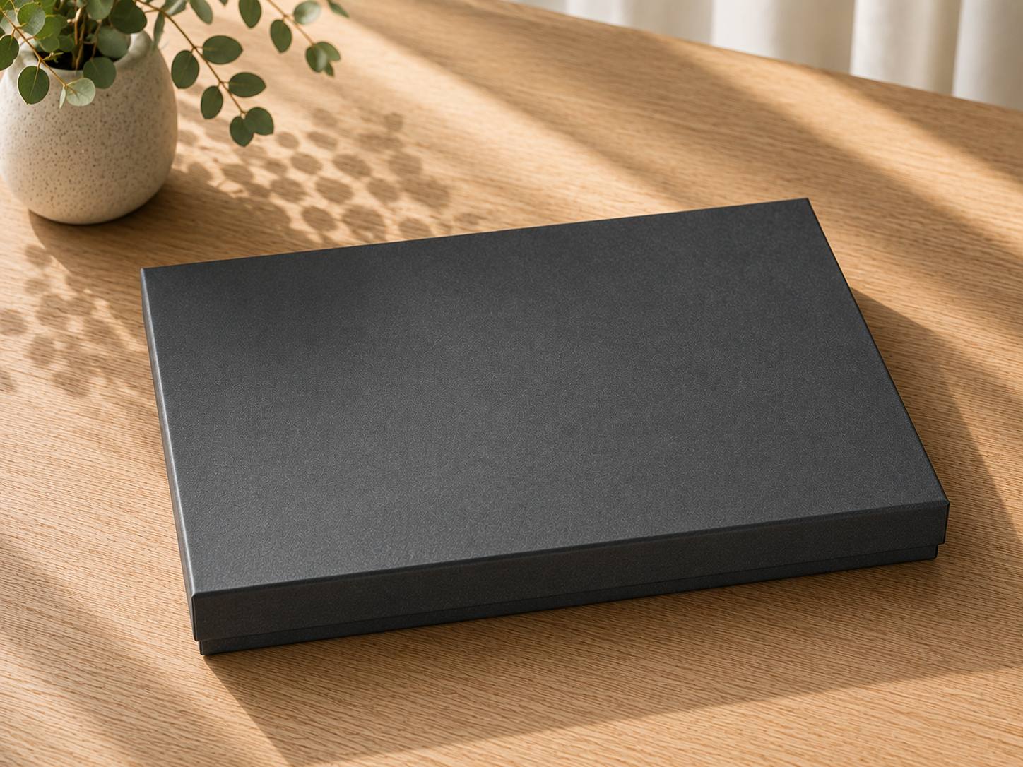 Eine geschlossene, rechteckige Geschenkbox „Pure A4“ von Artoz Papier AG in elegantem Schwarz steht auf einem hellen Holztisch neben einer Pflanze in einer kleinen Vase. Sonnenlicht wirft sanfte Schatten auf die Oberfläche und unterstreicht die edle Geschenkverpackung dieser exquisiten Geschenkbox „Pure A4“.