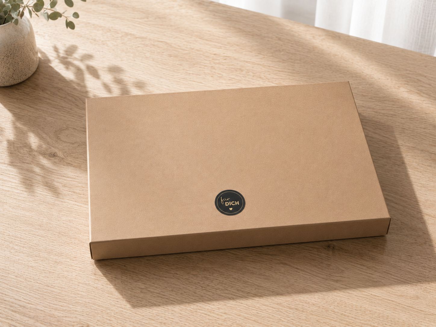Eine geschlossene, flache, braune „Geschenk-Box Pure A4“ von Artoz Papier AG mit einem kleinen schwarzen Aufkleber steht auf einer hellen Holzfläche neben einer Pflanze und zarten weißen Vorhängen und präsentiert sich als edle Geschenkverpackung für jeden Anlass.