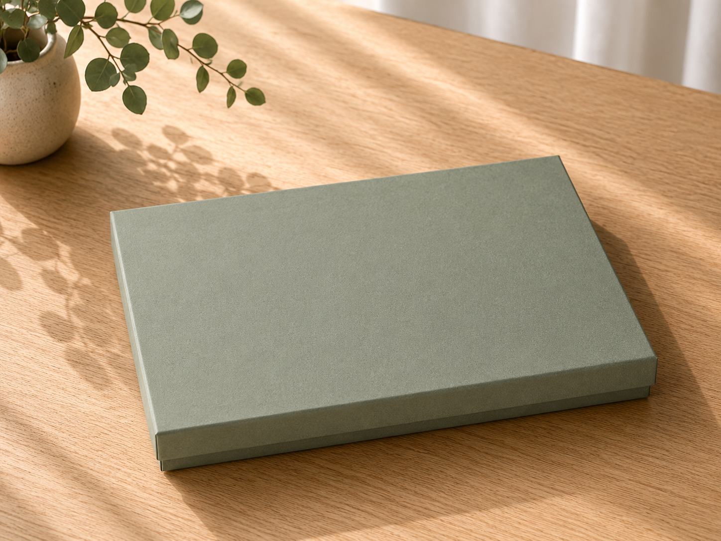 Eine geschlossene, rechteckige, grüne Geschenkbox „Pure A4“ von Artoz Papier AG steht auf einem hellen Holztisch, während das Sonnenlicht in der Nähe zarte Pflanzen-Schatten wirft. Diese edle Geschenkverpackung verleiht jedem Ambiente Eleganz.