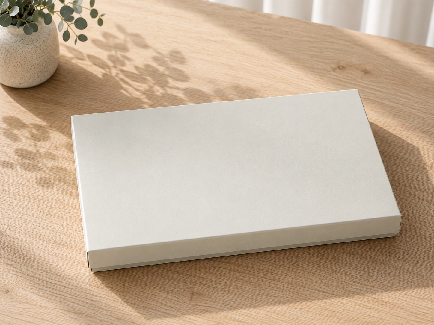Eine geschlossene, schlichte, rechteckige, weiße Geschenkbox „Pure A4“ von Artoz Papier AG steht auf einem hellen Holztisch neben einer kleinen Topfpflanze. Sonnenlicht wirft sanfte Schatten auf die Oberfläche und unterstreicht so die elegante Geschenkverpackung und ihre zurückhaltende Raffinesse.