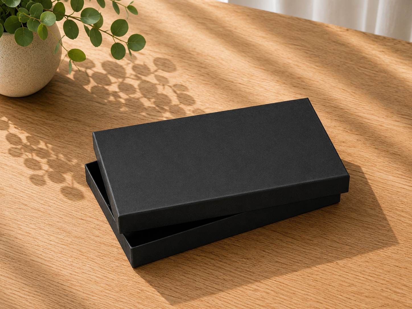 Sur une table en bois se trouve une « boîte cadeau Pure A5 » noire de la marque Artoz Papier AG, dont le couvercle est légèrement entrouvert, tandis que la lumière du soleil et les plantes projettent des ombres qui dansent sur la surface.