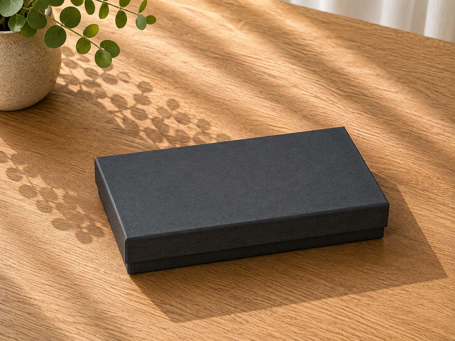 Sur une table en bois se trouve un coffret cadeau noir rectangulaire fermé, modèle « Pure A5 » de la marque Artoz Papier AG. La lumière du soleil projette les ombres d'une plante en pot aux feuilles luxuriantes située à proximité, créant des motifs sur le plateau de la table et mettant en valeur cet emballage raffiné.