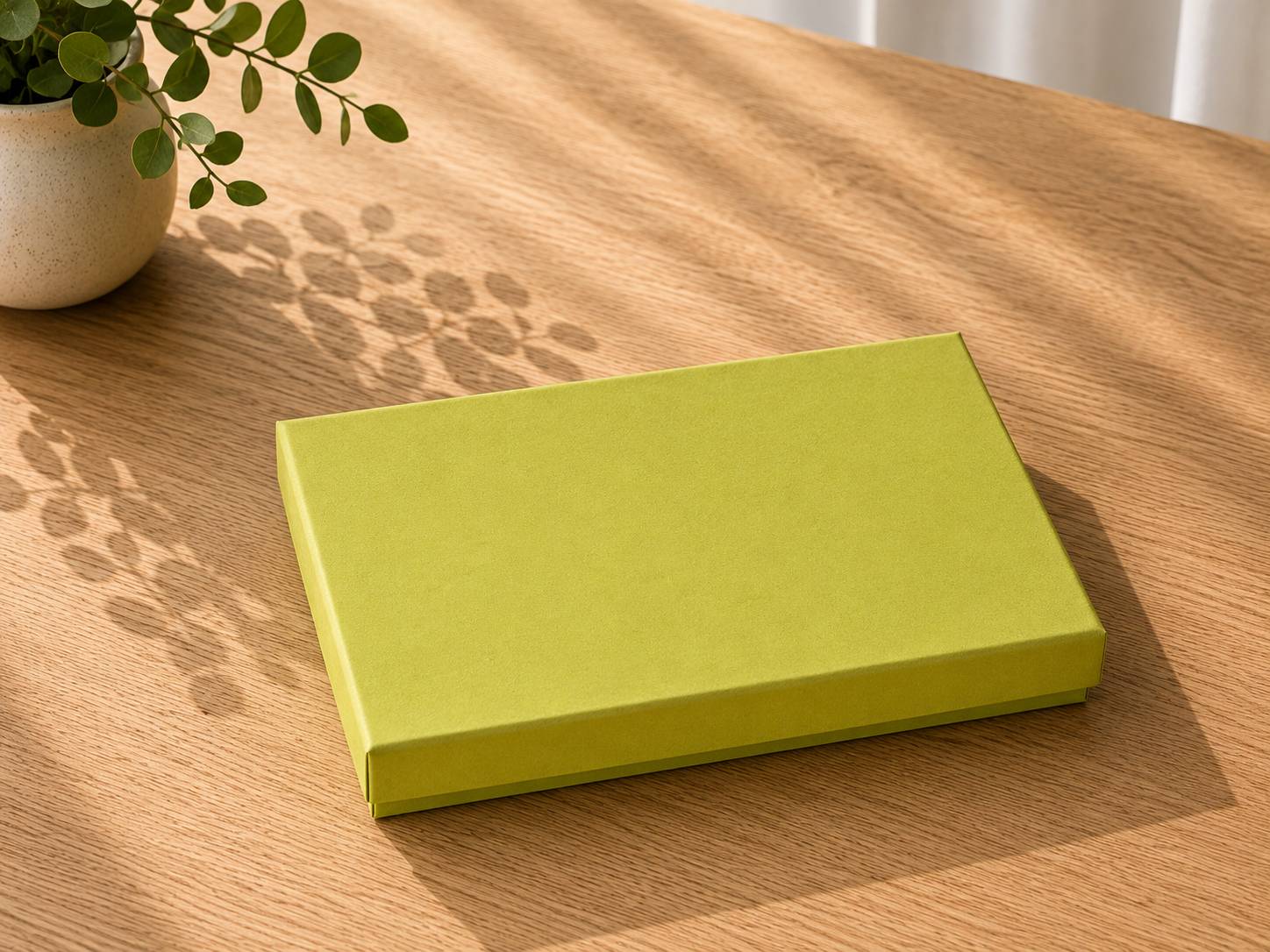 Eine geschlossene, rechteckige Geschenkbox „Pure A5“ in Grün von Artoz Papier AG steht auf einem Holztisch. Das Sonnenlicht wirft blattförmige Schatten einer nahe stehenden Zimmerpflanze auf die Tischplatte.