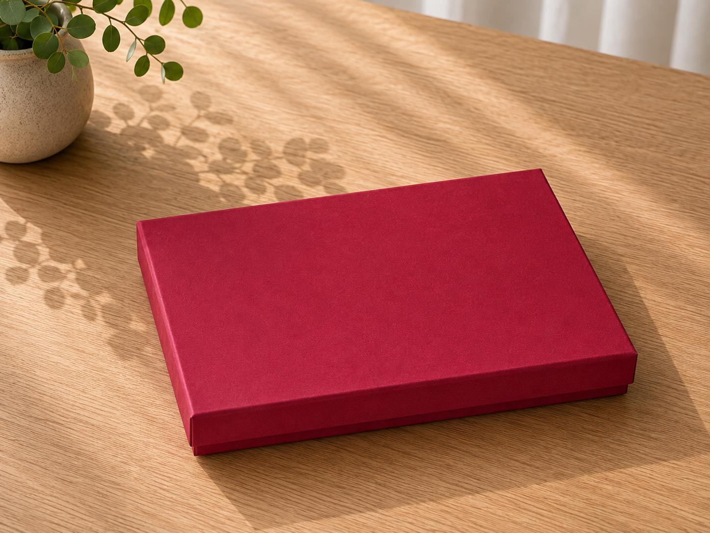 Une boîte-cadeau « Pure A5 » de la marque Artoz Papier AG, fermée, plate, rectangulaire et rouge, est posée sur une table en bois à côté d’une plante en pot ; la lumière du soleil pénètre dans la pièce et projette de délicates ombres de feuilles sur la surface. Cette solution d'emballage raffinée confère à la scène une touche d'élégance.