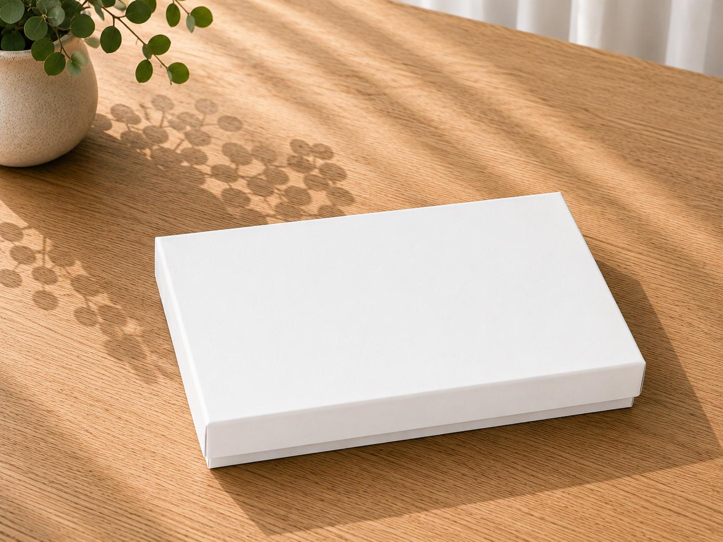 Auf einem Holztisch steht eine geschlossene, schlichte weiße Geschenkbox „Pure A5“ von Artoz Papier AG, auf die das Sonnenlicht Schatten von einer nahegelegenen Topfpflanze wirft.