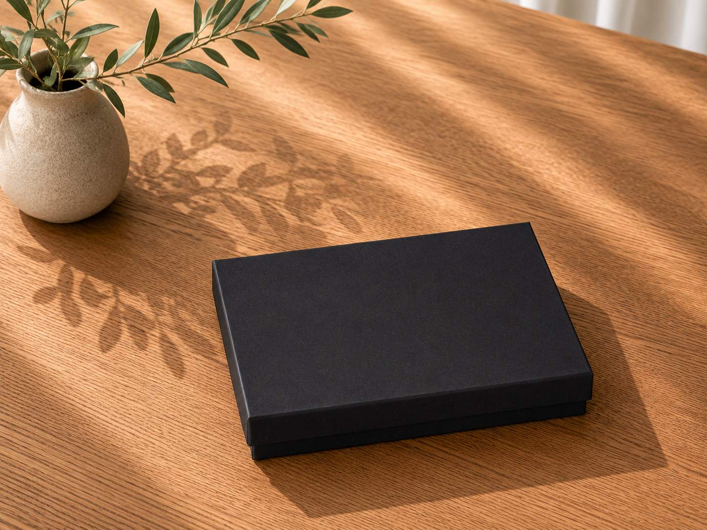 Auf einem Holztisch steht eine geschlossene schwarze „Geschenk-Box Pure A6“ von Artoz Papier AG neben einer beigen Vase mit grünen Blättern. Das Sonnenlicht wirft Blattschatten über den Tisch.