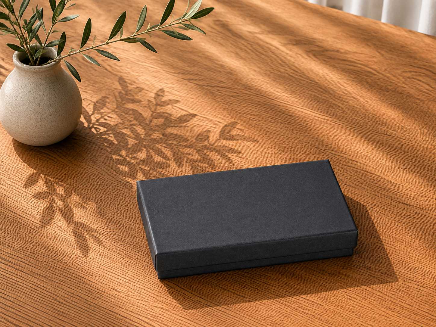 Eine geschlossene schwarze Geschenkbox „Pure A6“ der Artoz Papier AG steht auf einem Holztisch neben einer beigen Vase mit grünen, blättrigen Zweigen und wirft im natürlichen Sonnenlicht Schatten.