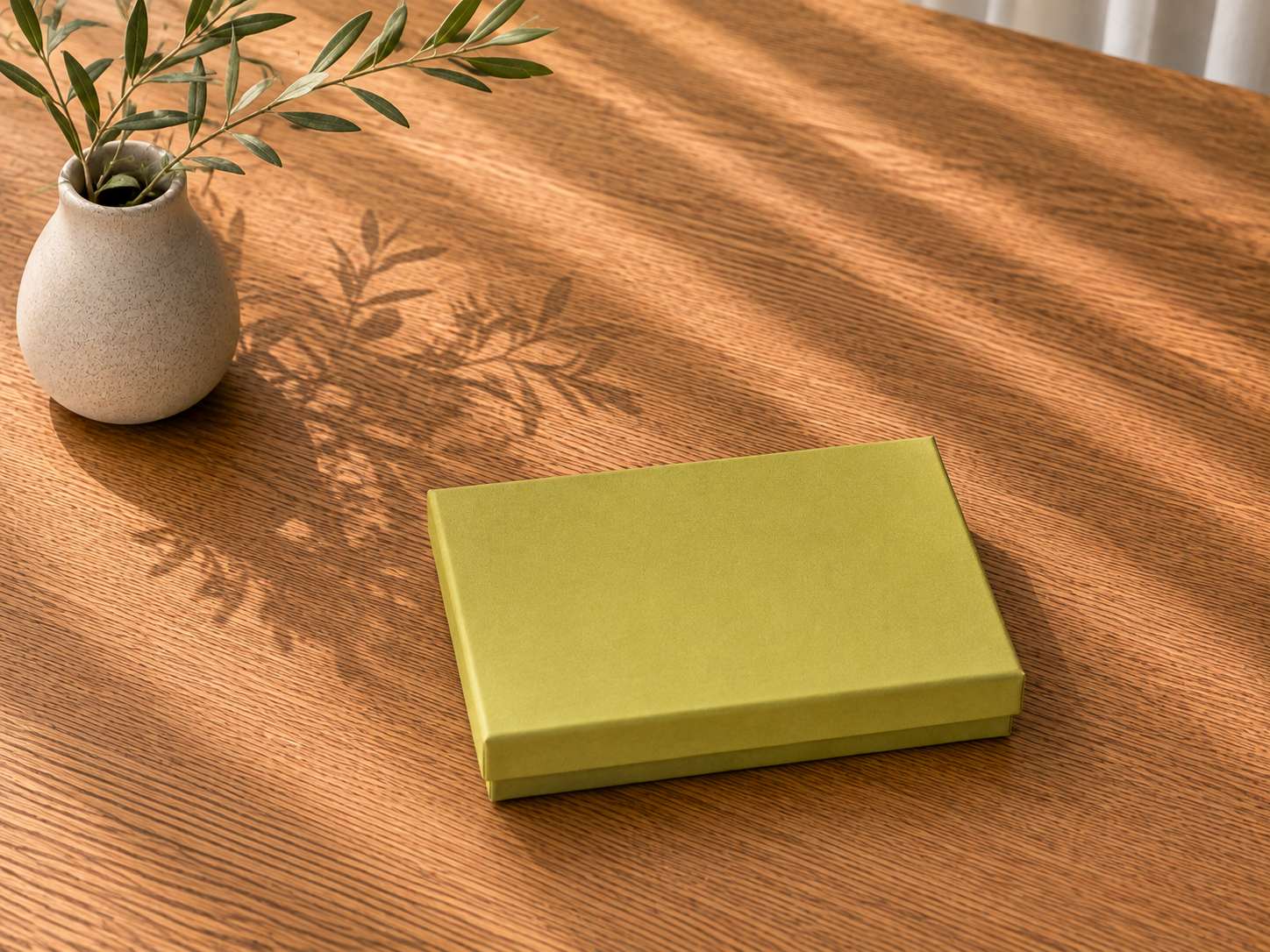 Eine grüne Geschenkbox „Pure A6“ der Artoz Papier AG steht geschlossen auf einem Holztisch neben einer kleinen Vase mit grünen Blättern, während Sonnenlicht und Schatten Muster über die Szene werfen.