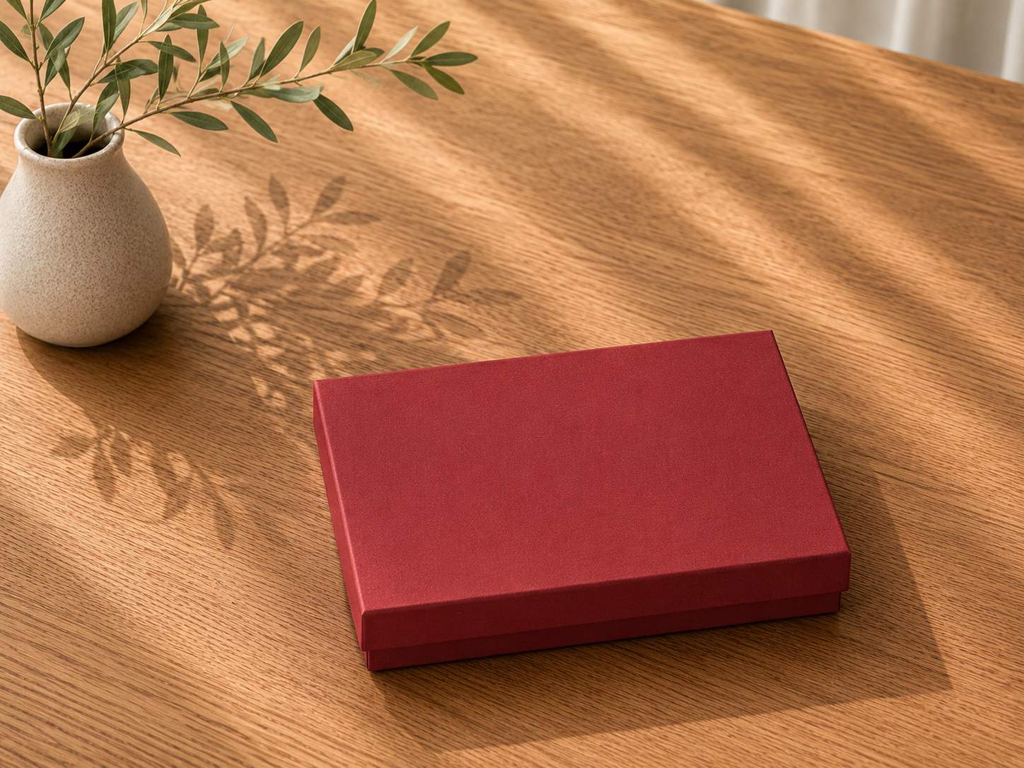 Eine „Geschenk-Box Pure“ im A6-Format in Rot von Artoz Papier AG steht geschlossen auf einem Holztisch neben einer kleinen weißen Vase mit einigen grünen Zweigen. Sanftes Sonnenlicht wirft weiche Schatten auf den Tisch.