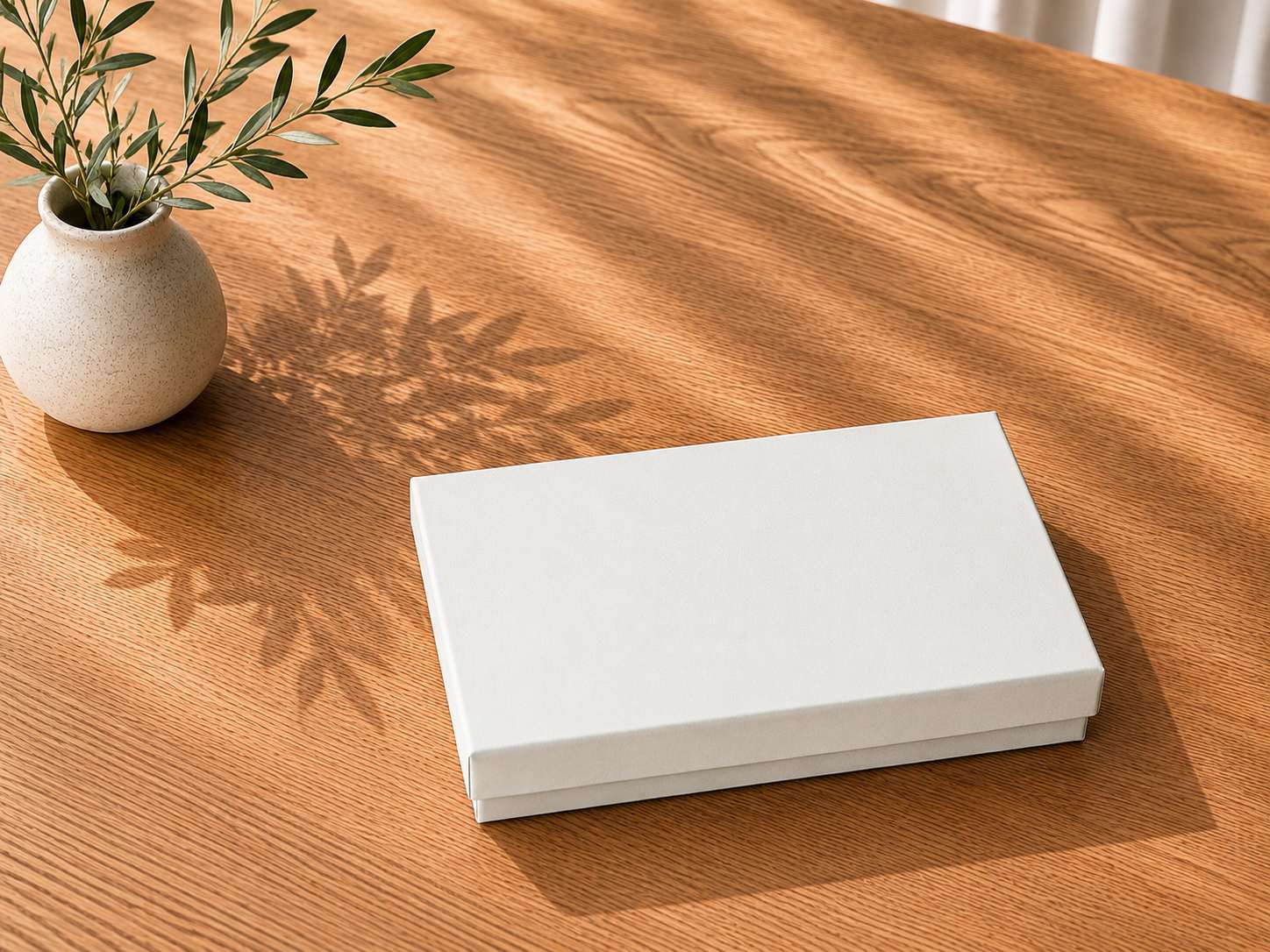 Eine geschlossene, schlichte weiße Geschenkbox „Pure A6“ in verschiedenen Farben von Artoz Papier AG steht auf einem hellen Holztisch neben einer kleinen runden Vase mit Olivenzweigen. Das Sonnenlicht wirft sanfte Schatten auf die Tischplatte und bringt die edle Verpackung dieser eleganten Schachtel besonders gut zur Geltung.