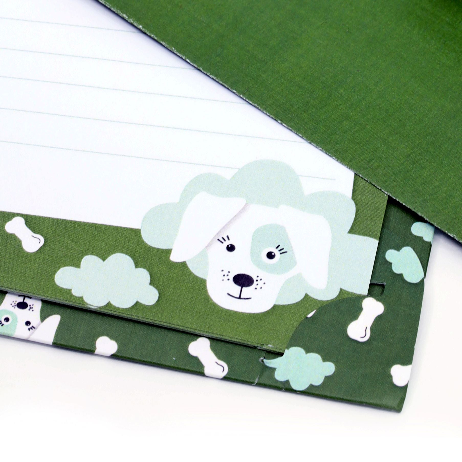 Gros plan sur le coffret de papier à lettres Kinder « Hund » de la marque Artoz Papier AG, de couleur verte, orné d'un adorable chien blanc, d'os et de nuages sur les bords. Certaines feuilles de ce coffret de papier à lettres pour enfants sont lignées et soigneusement empilées.