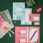 Une composition « flat lay » du coffret d'écriture pour enfants « Aliens » d'Artoz Papier AG, comprenant des cahiers illustrés d'animaux de dessins animés verts et roses, des cartes à lignes, un crayon, des petits cadeaux emballés dans du papier coloré et ornés d'un ruban, des étiquettes cadeaux rouges et une petite plante, le tout sur un fond vert.