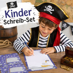 Un enfant souriant, déguisé en pirate, écrit sur du papier à lettres « Piraten » de la marque Artoz Papier AG, issu du coffret d'écriture pour enfants, assis à un bureau orné d'un coffre au trésor, de pièces de monnaie et d'un tonneau. Au-dessus, on peut lire « Kinder Schreib-Set », accompagné d'un motif représentant un drapeau de pirate.