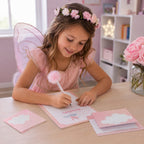 Une petite fille vêtue d'un costume de fée sourit tandis qu'elle s'entraîne à l'écriture créative avec le kit d'écriture pour enfants « Wolke » d'Artoz Papier AG, assise à son bureau dans une chambre aux tons pastel.