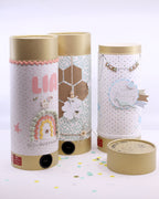 Das "Tischbombe ohne Feuer - DIY 3er Set" der Artoz Papier AG besteht aus drei aufrecht stehenden zylindrischen Schachteln mit goldenen Deckeln, pastellfarbenen Motiven, festlichen Botschaften und Ornamenten - perfekt für kinderfreundliche Feste. Buntes Konfetti schmückt die weiße Fläche drumherum.