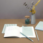 Ein Arbeitsbereich mit dem Artoz Papier AG Notizblock 3er Set farbig (100x180mm), einem offenen Notizbuch, einem blauen Bleistift, verschiedenen Stiften, einem gestreiften geschlossenen Notizbuch, einem Glas mit Büroklammern und einer Vase mit gelben und grünen Trockenblumen auf einem braunen Schreibtisch.