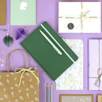 Le carnet de notes Artoz Papier AG A5 avec stylo vert gravé à ton nom, des crayons décoratifs, des pochettes cadeaux, des cadeaux emballés et différentes cartes de vœux sur fond violet.