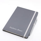 Un carnet A5 gris avec stylo de la marque Artoz Papier AG, muni d'un élastique blanc et d'une couverture personnalisable sur laquelle est imprimé le nom « Stefanie Riner » en lettres blanches.