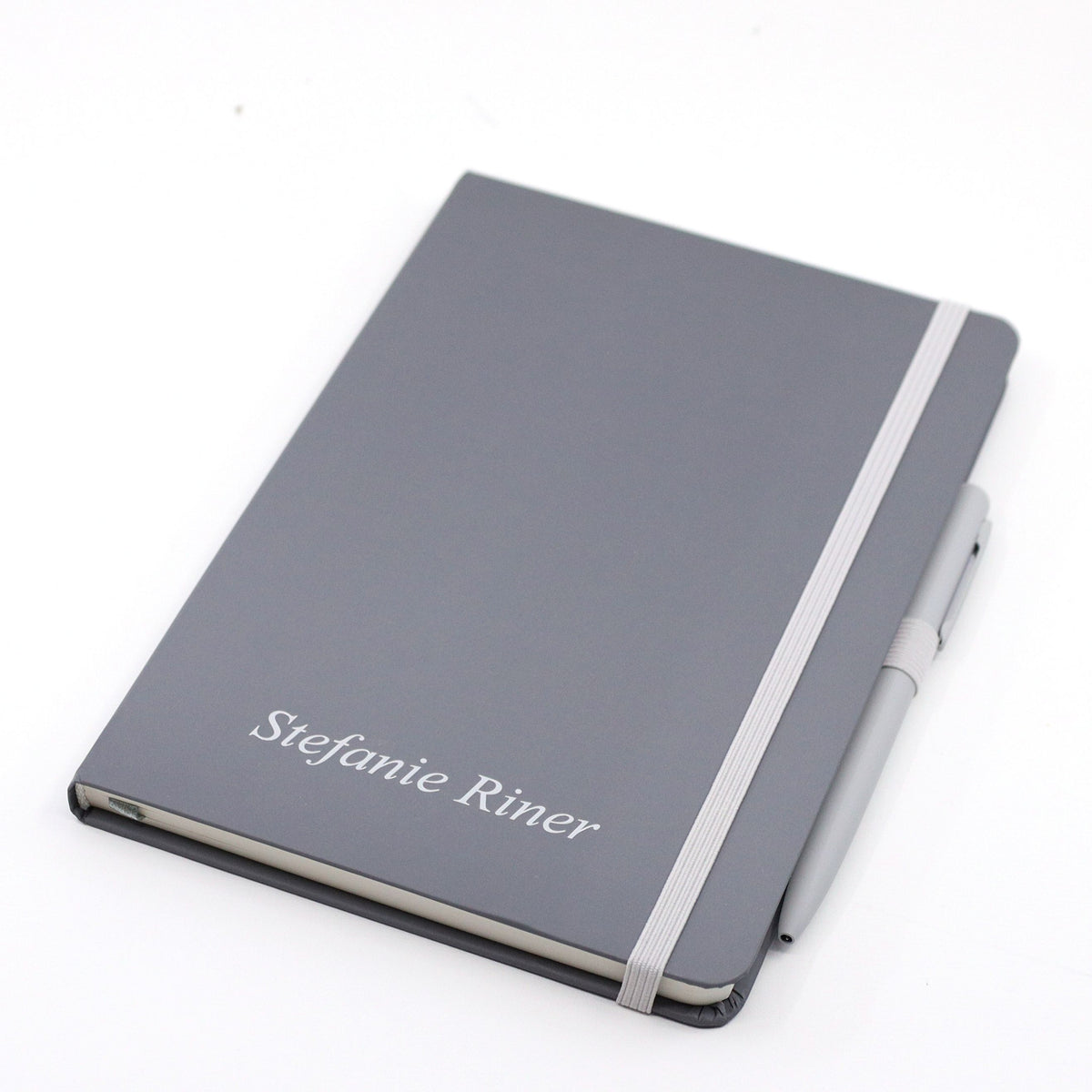 Un carnet A5 gris avec stylo de la marque Artoz Papier AG, muni d'un élastique blanc et d'une couverture personnalisable sur laquelle est imprimé le nom « Stefanie Riner » en lettres blanches.