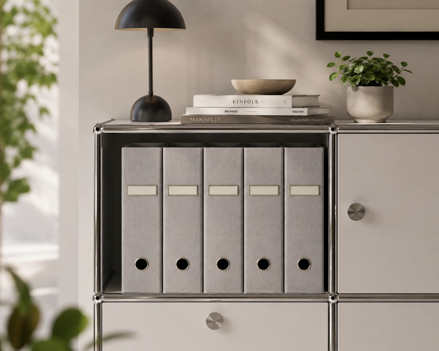 Cinq classeurs A4 gris de la marque Artoz Papier AG sont soigneusement rangés sur une étagère où se trouvent également une lampe noire, des livres empilés, un bol et une petite plante en pot. Cette composition minimaliste et bien éclairée est idéale pour votre bureau à domicile.