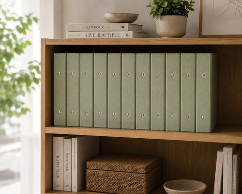 Une bibliothèque en bois accueille des classeurs A4 « schmal » d'Artoz Papier AG à l'aspect lin, des livres empilés sur l'étagère du haut et des paniers tressés en dessous — l'idéal pour un bureau à domicile bien rangé. Une plante à feuillage luxuriant vient agrémenter l'arrière-plan.