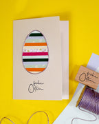 Der Stempel Frohe Ostern von Artoz Papier AG zeigt eine beige Osterkarte mit einem bunt gestreiften Eierausschnitt und dem Text "Frohe Ostern", dargestellt auf Gelb mit passendem Stempel und lila Faden, ideal für kreative Bastelarbeiten.