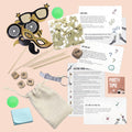 Das Artoz Papier AG Inhaltset für Tischbomben - Thema "Party" mit Fotorequisiten, Konfetti, Spielkarten, Holzwürfel, "Cheers to us!"-Schlüsselanhänger, Stoffbeutel, Haftnotizen und zwei grünen Hüpfkugeln auf rosa Hintergrund.