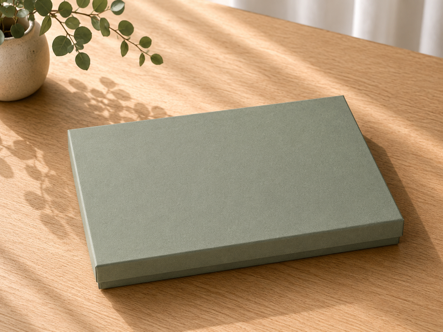 Eine geschlossene, rechteckige, grüne Geschenk-Box „Pure A6“ von Artoz Papier AG steht auf einem Holztisch neben einer Topfpflanze, wobei das Sonnenlicht sanfte Schatten auf die Oberfläche wirft.