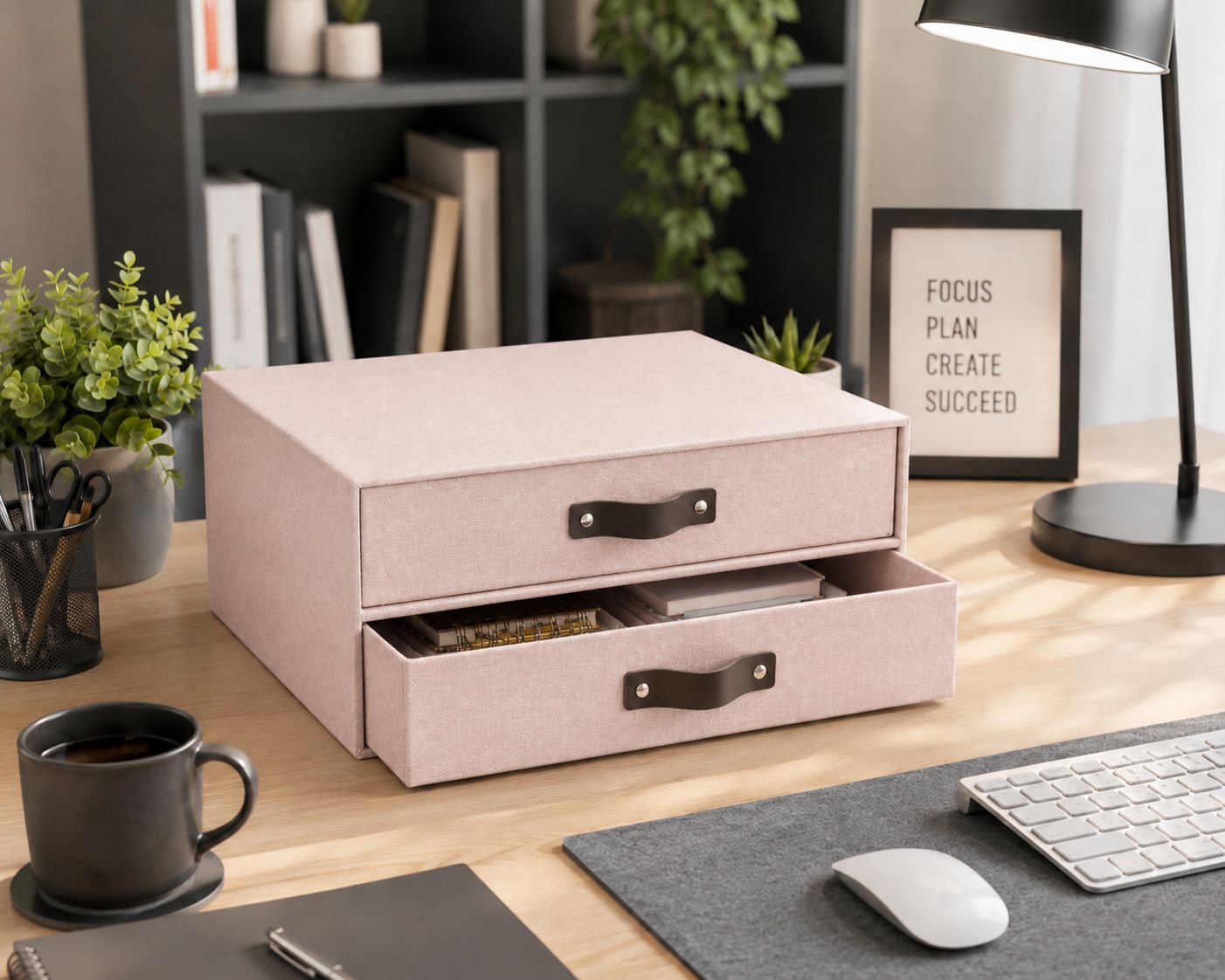 Auf einem aufgeräumten Schreibtisch steht eine rosa Schubladenbox von Artoz Papier AG in Leinenoptik, deren eine Schublade geöffnet ist, daneben eine Tastatur, eine Lampe, Pflanzen, ein Notizbuch und ein gerahmtes Schild mit der Aufschrift „FOCUS PLAN CREATE SUCCEED“.