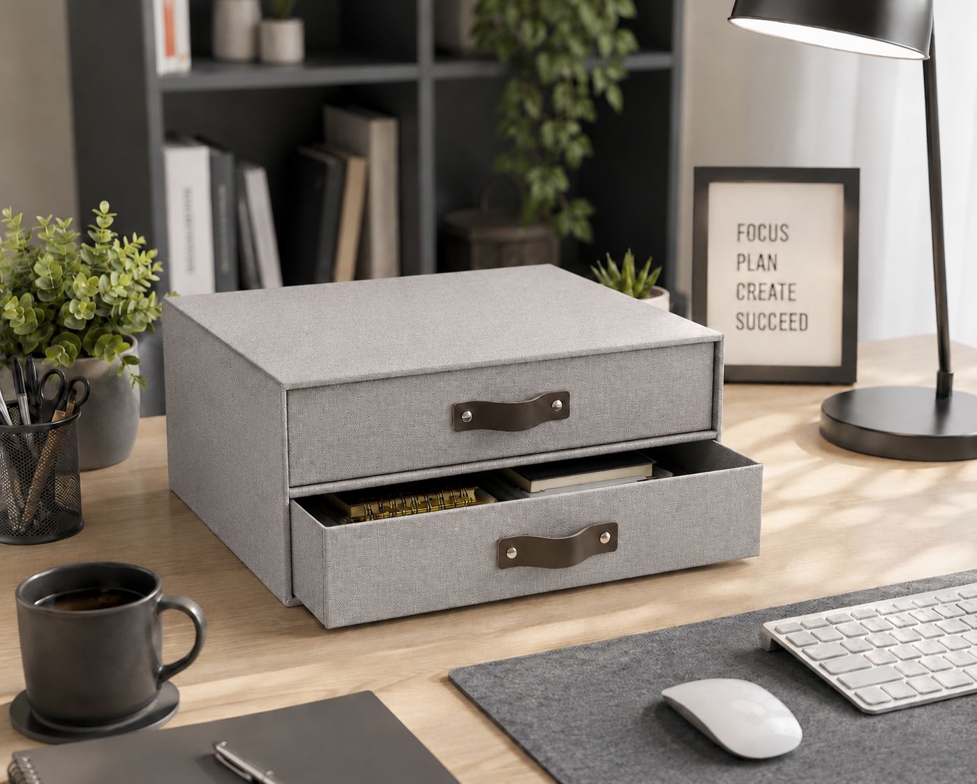 Ein moderner Schreibtisch mit einer Schubladenbox von Artoz Papier AG in grauer Leinenoptik, bei der eine Schublade geöffnet ist, sowie einer Tastatur, einem Notebook, einer Kaffeetasse, einer Lampe, Pflanzen und einem gerahmten Motivationszitat im Hintergrund – für eine perfekte Büroorganisation.