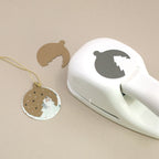Ein Papierstanzer „M“ der Artoz Papier AG mit einem Weihnachtskugel-Motiv ist neben einem ausgestanzten Ausschnitt und einem fertigen Anhänger zu sehen, der mit einer goldenen Schnur, Sternen und einem Baum verziert ist. Auf einem beigen Untergrund angeordnet, bieten diese Artikel festliche Inspiration für Bastel- und Kartenprojekte.