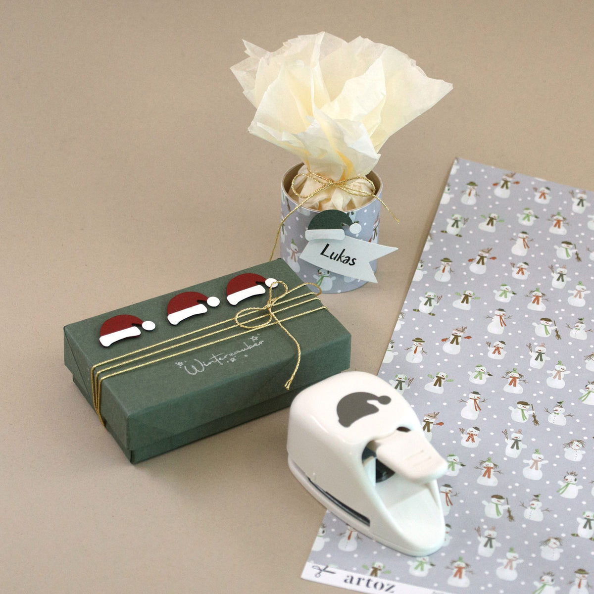 Un coffret cadeau vert orné de bonnets de Père Noël, une tasse décorée remplie de papier de soie, la perforatrice « Stanzer S Mütze » d'Artoz Papier AG et du papier d'emballage festif à motifs de pingouins — idéal pour la fabrication de cartes — sont disposés sur un support beige.
