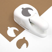 Une perforatrice artisanale « Stanzer M Weihnachtskugel » de la marque Artoz Papier AG repose sur du papier marron et blanc, découpant des formes circulaires ornées d'un motif de sapin dentelé au centre — idéale pour la fabrication de cartes ou pour ajouter une touche festive à vos projets.