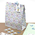 Eine geblümte Geschenktüte mit einem DANKE-Anhänger, verziert mit einem blauen Schmetterling und Papierblumen. In der Nähe befindet sich der Artoz Sticker "Schmetterling transparent" von Artoz Papier AG, sowie Papierblumen-Verzierungen und pastellfarbenes Schmetterlings-Briefpapier.
