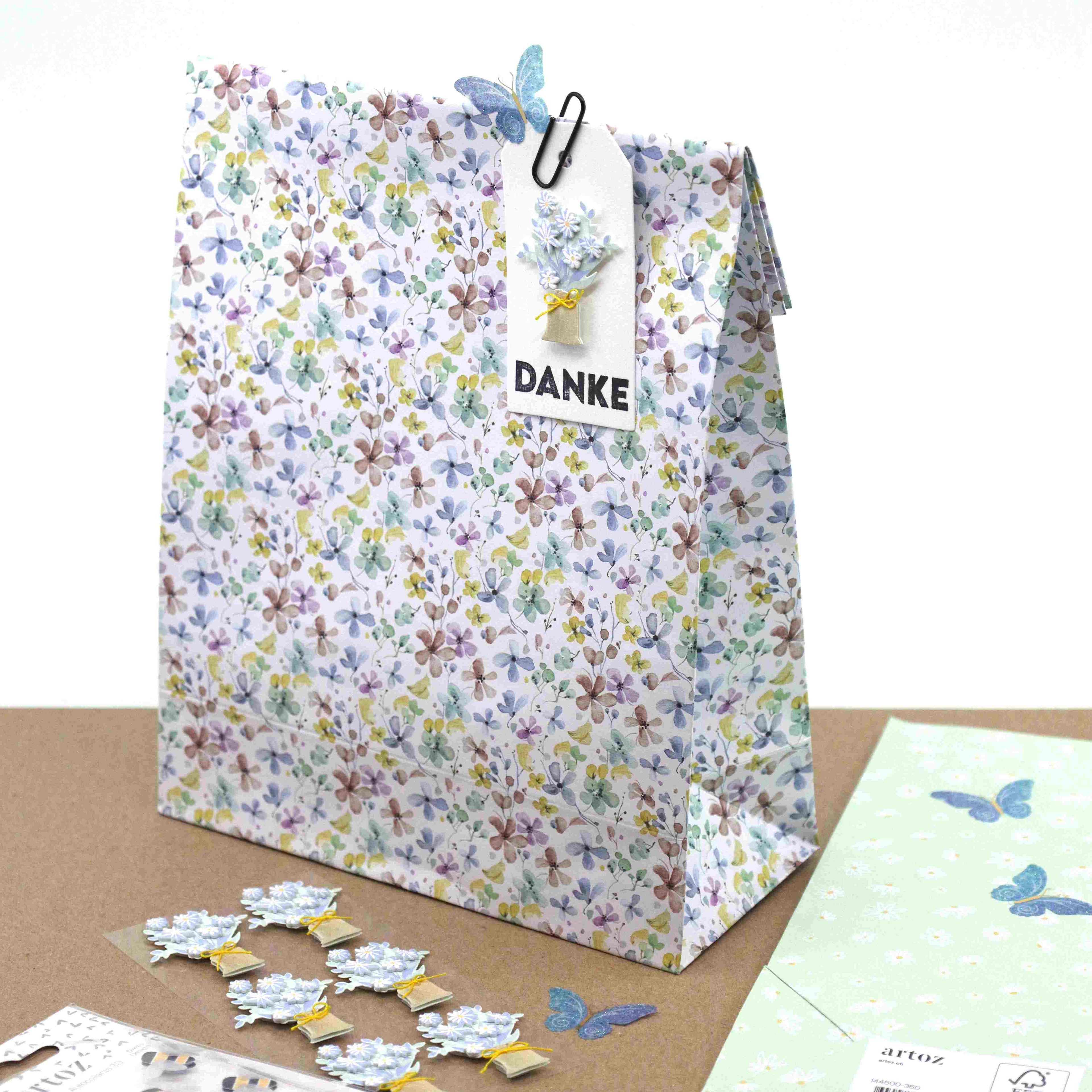 Eine geblümte Geschenktüte mit einem DANKE-Anhänger, verziert mit einem blauen Schmetterling und Papierblumen. In der Nähe befindet sich der Artoz Sticker "Schmetterling transparent" von Artoz Papier AG, sowie Papierblumen-Verzierungen und pastellfarbenes Schmetterlings-Briefpapier.