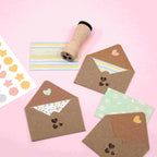 Ein flacher Bogen mit drei offenen braunen Umschlägen mit gemusterten Einlagen, einer gestreiften Karte, einem Stempel mit Holzgriff und einem Artoz Papier AG Artoz Sticker "Mini Icons" auf einem rosa Hintergrund.