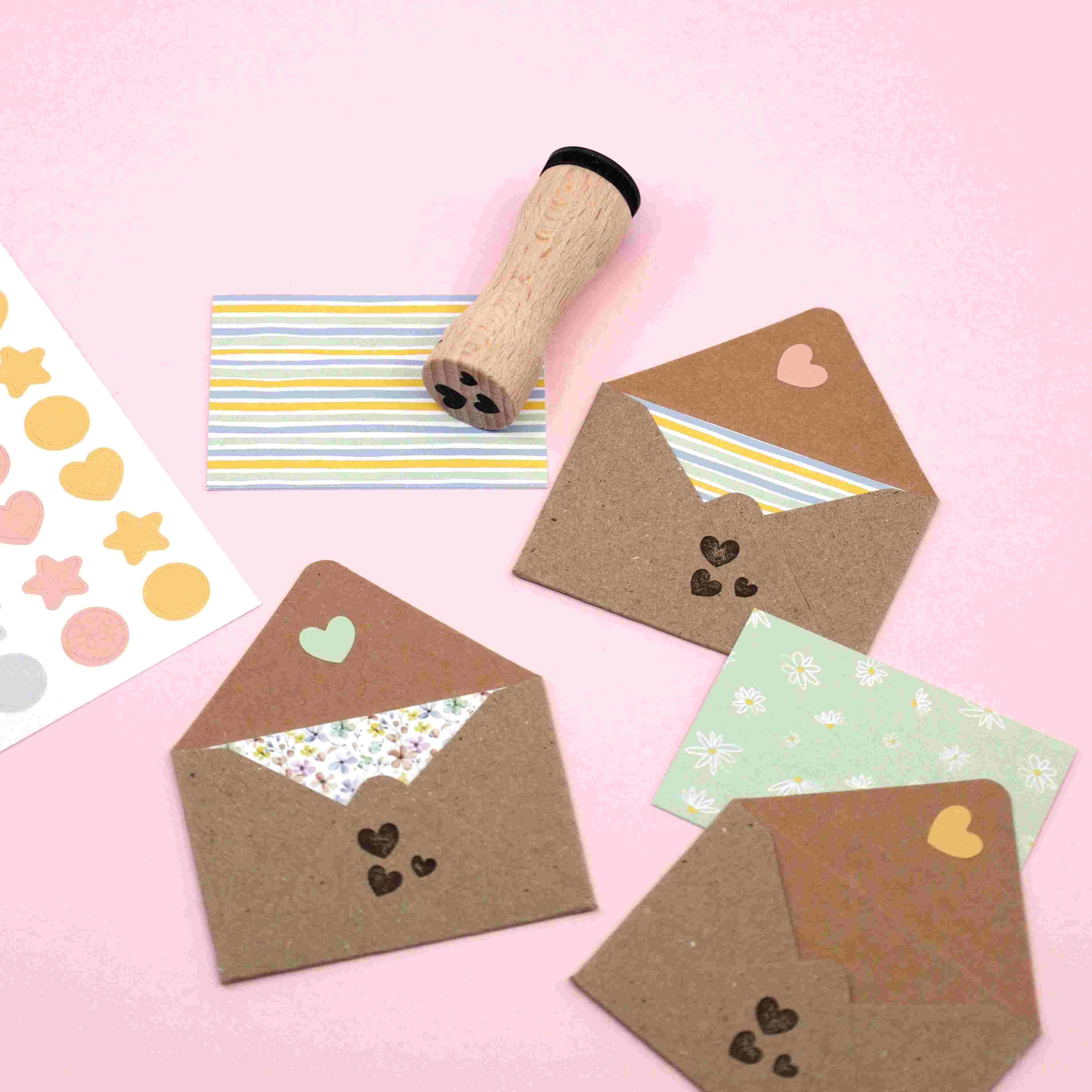 Ein flacher Bogen mit drei offenen braunen Umschlägen mit gemusterten Einlagen, einer gestreiften Karte, einem Stempel mit Holzgriff und einem Artoz Papier AG Artoz Sticker "Mini Icons" auf einem rosa Hintergrund.