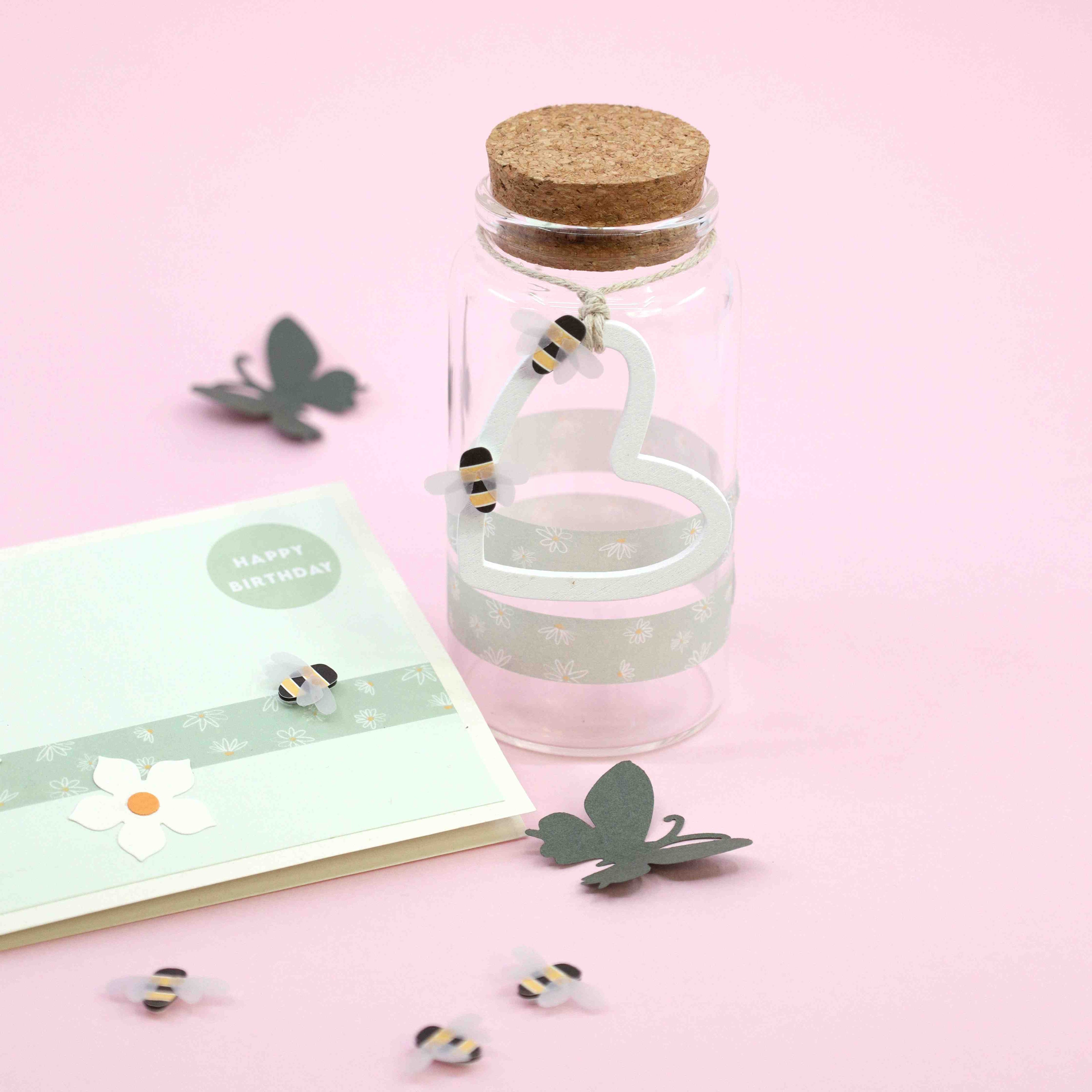 Un bocal en verre avec un couvercle en liège se trouve à côté d'une carte d'anniversaire pastel décorée avec l'autocollant Artoz 3D Sticker "Biene" d'Artoz Papier AG, des fleurs en papier et des papillons éparpillés sur un fond rose.