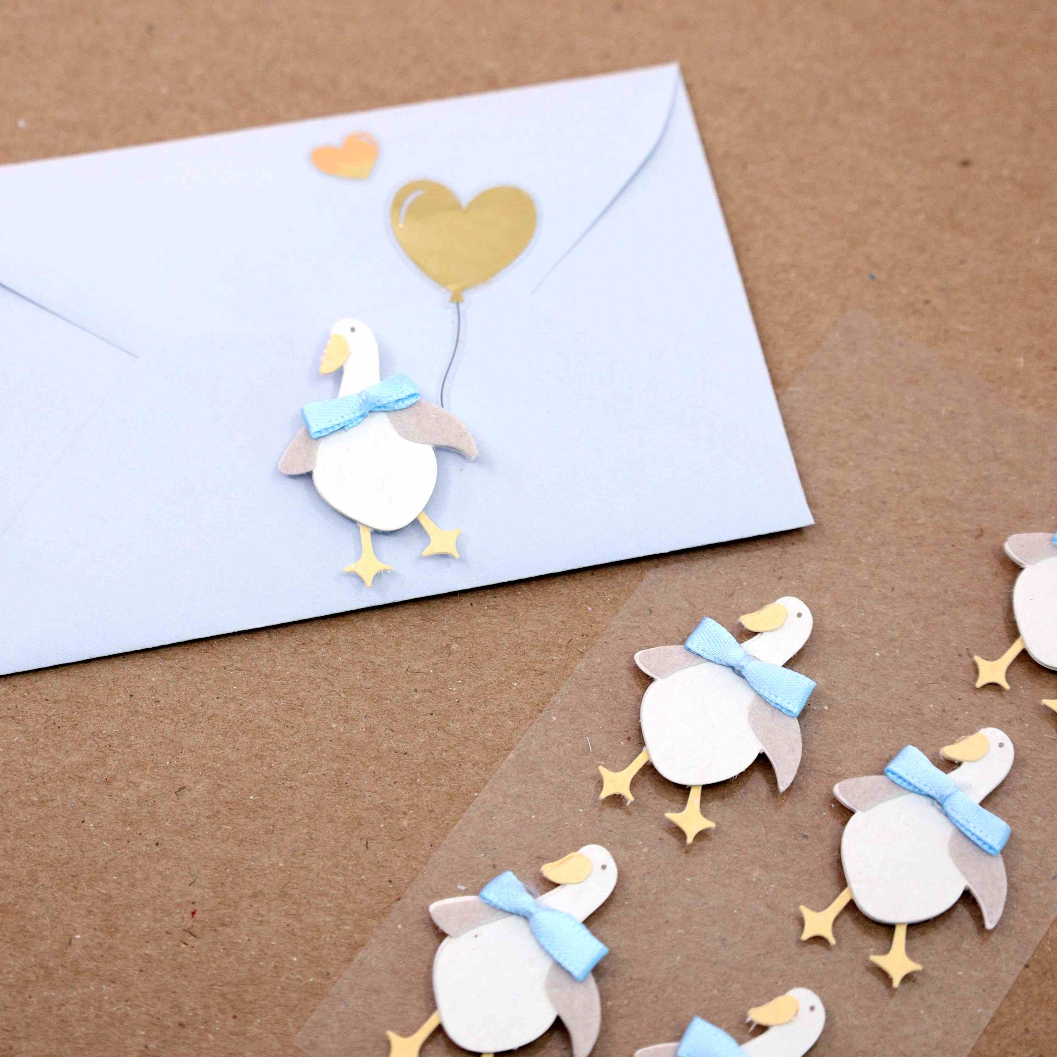 Une enveloppe blanche, décorée avec les Artoz 3D Sticker Gans d'Artoz Papier AG, présente une oie tenant un ballon à cœur d'or et un petit cœur d'or, associés à des découpes d'oies assorties à nœuds bleus, idéales pour les projets de bricolage de design suisse.