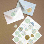 Zwei verzierte Briefumschläge und ein Artoz Sticker Happy Birthday Bogen der Artoz Papier AG mit Herz- und Pastellmotiven werden auf einer braunen Fläche präsentiert - ideal für Bastelprojekte oder als charmante Geburtstagsdeko.