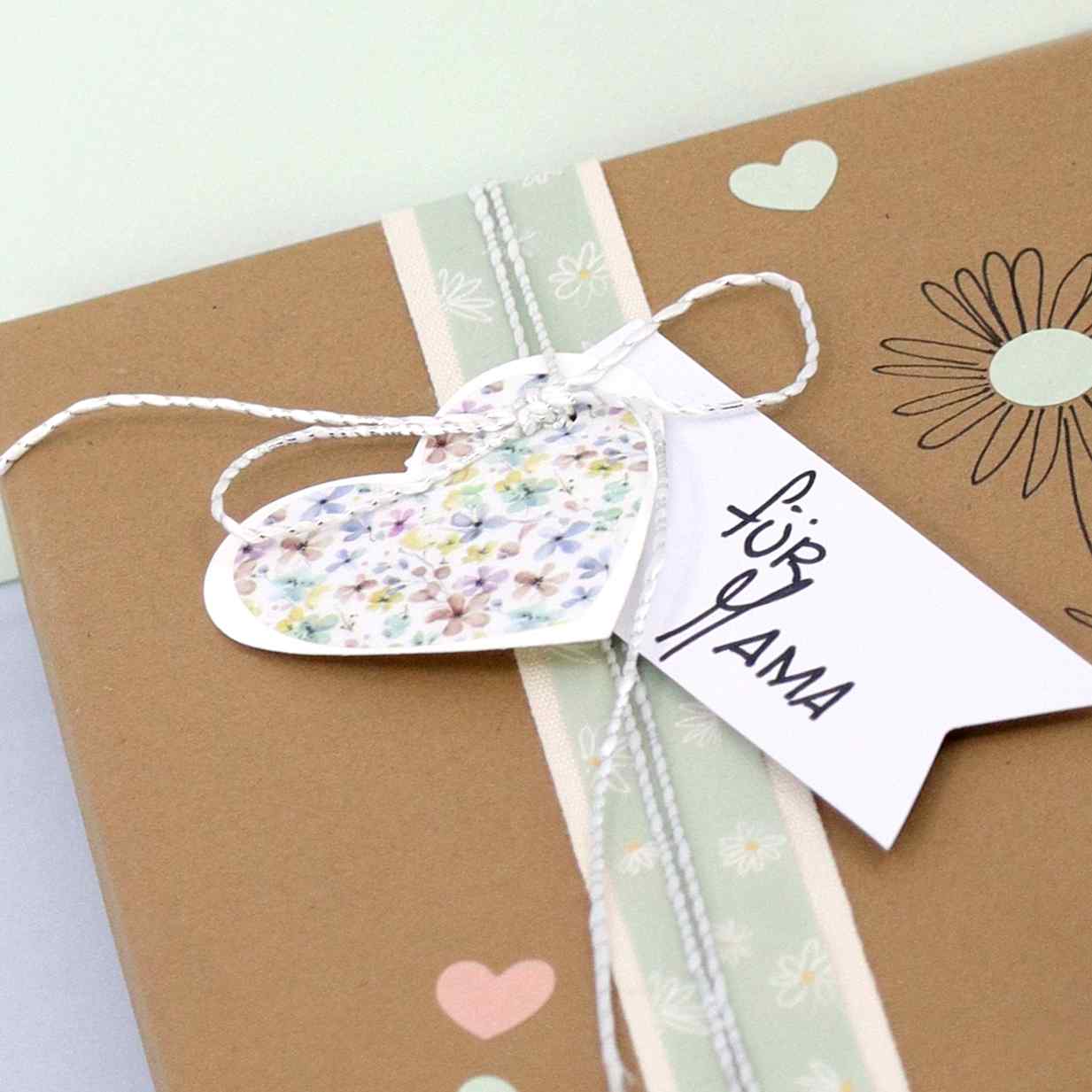 Eine braune Geschenkbox mit floralem Muster, umwickelt mit grünem und weißem Band, verziert mit einem Artoz Sticker "Herz transparent" der Artoz Papier AG, einem floralen Herzanhänger und einer "für MAMA"-Karte - ideales Bastelzubehör für Geschenke.