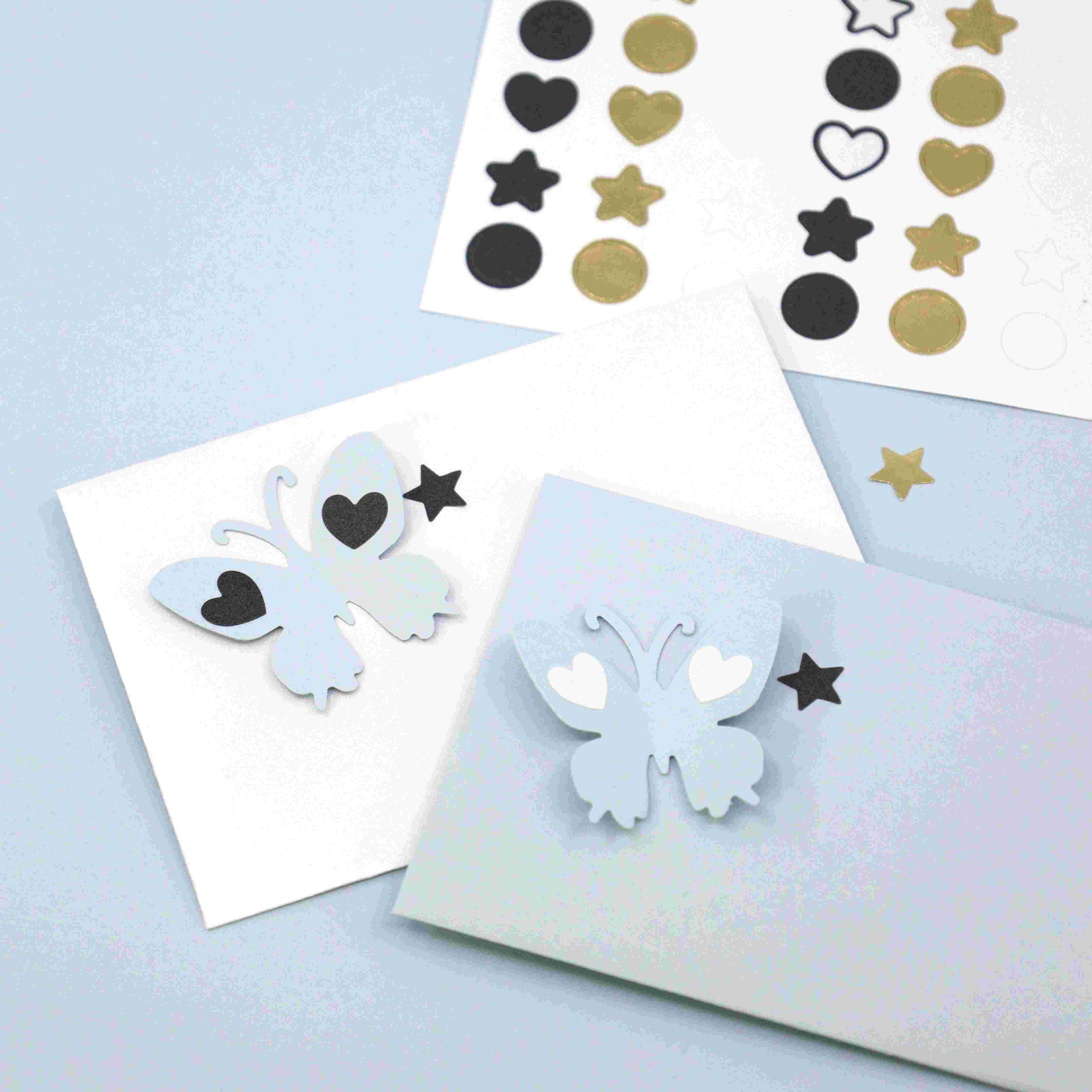 Deux cartes blanches sont ornées de papillons en papier 3D et de cœurs, étoiles et cercles Artoz Papier AG Sticker. Les feuilles d'autocollants "Mini Icons Basic" d'Artoz Papier AG sont visibles - idéales pour décorer des cadeaux sur une surface bleu clair.