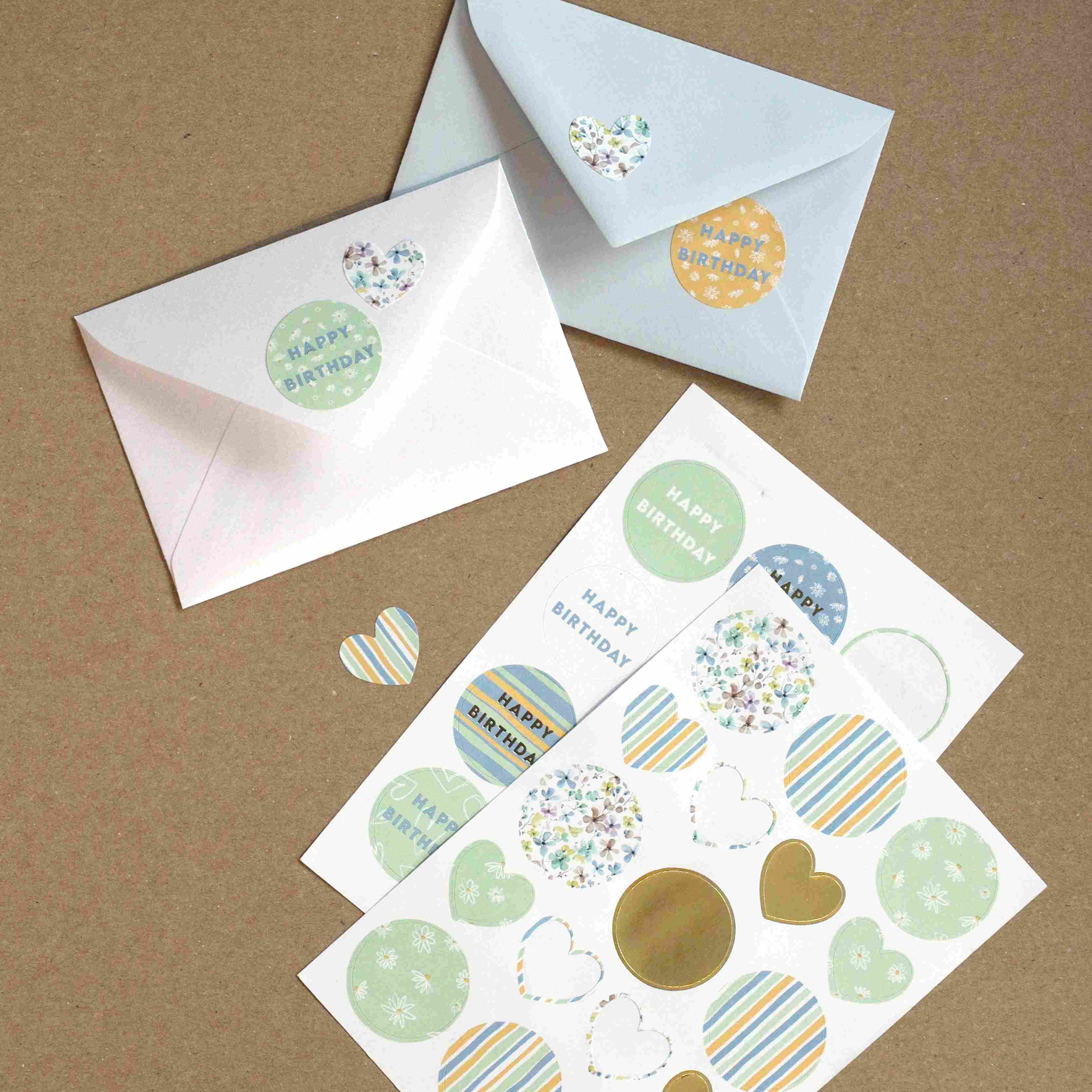 Deux enveloppes, l'une blanche et l'autre bleu clair, sont fermées par des autocollants Artoz Papier AG ronds et en forme de cœur. A proximité se trouve la feuille d'autocollants Artoz Sticker "Mixed Frühling" avec des motifs pastel de joyeux anniversaire, parfaits pour le Bastelzubehör ou les bricolages de printemps.