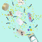 Disperse sur un fond bleu clair, la « Tischbombe ohne Feuer » - Schulstart d’Artoz Papier AG propose un assortiment de petits jouets, de bonbons, de confettis et de pièces de jeu — notamment un jeu de morpion, des jetons emoji, un tube sur le thème des animaux et une boîte ronde — pour offrir une alternative adaptée aux enfants à la bombe de table classique pour la rentrée scolaire, ainsi qu’une option réutilisable.