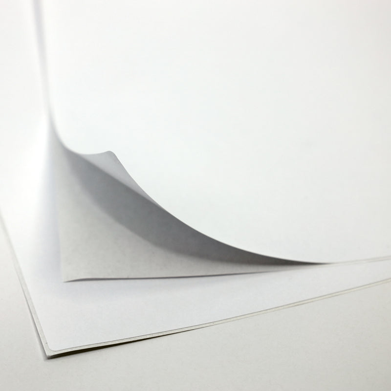 Gros plan sur une pile d'Artoz Papier AG A4 Klebefolie zum selber bedrucken - für Tischbombe DIY, la feuille supérieure étant partiellement soulevée pour révéler les couches inférieures, sur un fond blanc uni.