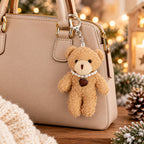 Eine beige Handtasche, verziert mit dem Artoz Papier AG Teddy-Anhänger – Plüschbär mit Herzkette, der eine Perlenkette und einen braunen Herzanhänger trägt, liegt auf einer Holzunterlage, umgeben von festlicher Winterdekoration – und ist damit das perfekte Wintergeschenk.
