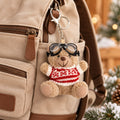 Un pendentif en forme d'ours en peluche portant des lunettes de ski, fabriqué par Artoz Papier AG, est accroché à la fermeture éclair d'un sac à dos beige. Le petit ours en peluche porte un pull rouge et blanc ainsi que des lunettes de ski. En arrière-plan, des lumières floues et des brindilles vertes créent une ambiance chaleureuse : un charmant cadeau d'hiver !.