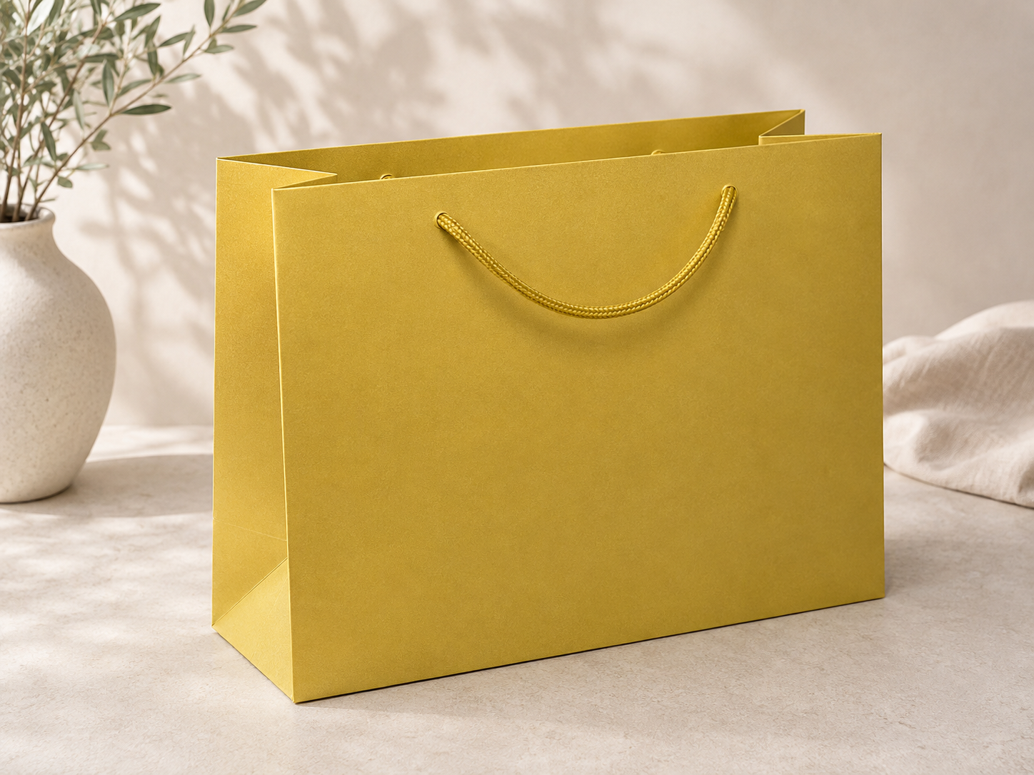 Eine Geschenktasche „Pure“ der Marke Artoz Papier AG in Größe L in Gelb mit Kordelgriffen steht auf einer hellen Steinoberfläche. Im Hintergrund ist eine weiße Vase mit grünen Blättern zu sehen, auf die sanftes, natürliches Licht fällt und sanfte Schatten wirft.